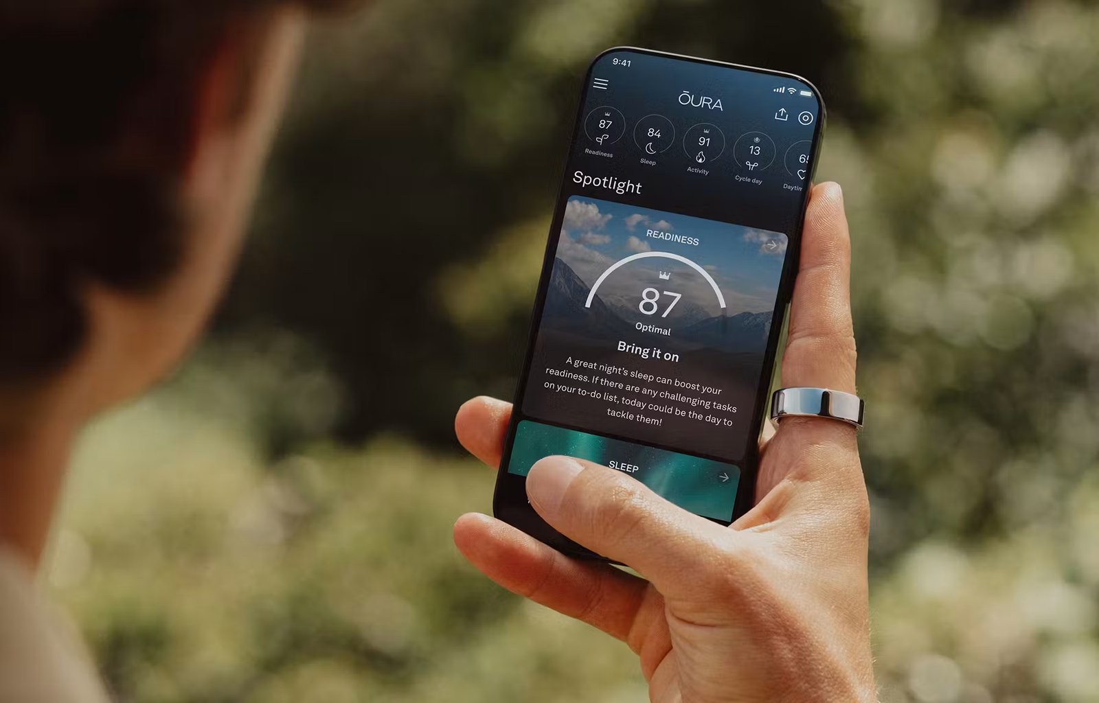 A person views the Oura app on their smartphone while wearing an Oura ring on their right index finger